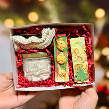 Cargar imagen en el visor de la galería, Caja navideña artesanal con whipped body butter de 2.5 oz, dos jabones decorados y bolsa de sisal sobre relleno rojo.