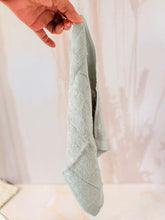 Cargar imagen en el visor de la galería, Toallita sostenida: Mano sosteniendo una toallita facial orgánica de color gris claro, mostrando caída natural y suavidad sobre fondo neutro.