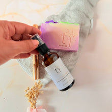 Cargar imagen en el visor de la galería, Spray sanitizante artesanal Manitas Limpias con aroma a lavanda, colocando junto a una barra barras de jabón de Lavanda, una toalla gris y un lapicero de unas sobre un fondo claro y florecillas de etatie secas Producto de Tt atelier.