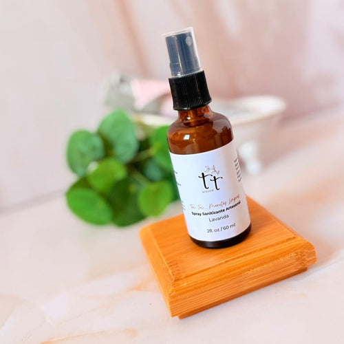 Spray sanitizante artesanal Manitas Limpias con lavanda sobre base de madera, con hojas verdes al fondo y fondo claro. Producto de Tt atelier